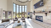 Located at 11245 West Rd in Houston, TX, this single-floor community space is a masterfully designed gathering area that blends elegance with comfort. The centerpiece of the space is a dramatic curved bay of floor-to-ceiling windows that floods the interior with abundant natural light while offering sweeping views of a beautifully landscaped courtyard and outdoor amenity areas. Soaring ceilings amplify the sense of grandeur throughout.

A stunning full-height natural stone fireplace with a rustic wood mantel serves as a striking architectural focal point, anchoring the main lounge area. Light wood-tone tile flooring runs throughout, complementing the neutral, contemporary color palette. The lounge features sophisticated seating arrangements, including tufted sofas and accent chairs, creating an inviting and well-appointed atmosphere.

The space flows seamlessly into a bright dining area with a round wooden table and navy accent chairs, and further into a well-equipped kitchenette. The kitchenette offers sage green cabinetry, open wood shelving, a beverage refrigerator, and modern appliances, making it ideal for communal use. Recessed lighting and large windows ensure every area of the space remains bright and welcoming throughout the day.