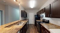 This well-appointed single-story, 3-bedroom, 2-bathroom home offers a thoughtful layout designed for comfortable everyday living. The galley-style kitchen is a functional centerpiece, featuring rich dark espresso cabinetry, granite-style countertops, a double stainless steel sink, and black stainless appliances including an electric range and refrigerator. Wood-look flooring runs throughout the kitchen, adding warmth and durability to the space. The kitchen opens toward a combined living and dining area, where a peninsula counter provides a natural transition between spaces. The living room is carpeted and anchored by a tiled fireplace, creating a welcoming focal point. A pendant light fixture marks the dining area, which flows seamlessly into the main living space. Each of the three bedrooms is finished with plush carpet, ceiling fans with light fixtures, and windows that allow natural light to fill the rooms. The two bathrooms are well-designed, each featuring dark wood vanities with granite-style countertops, wood-look flooring, and tiled tub-shower combinations. The primary bathroom benefits from added natural light through a window and an oversized mirror above the vanity. Crown molding and consistent neutral tones throughout contribute to a clean, cohesive aesthetic across the entire home.