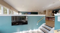 This single-floor community space is thoughtfully designed to serve as both a welcoming gathering area and a functional workspace. The interior is defined by a distinctive split-level layout, where a bold teal accent wall anchors the main lounge area, contrasted by crisp white trim and neutral taupe upper walls that lend the space a clean, polished aesthetic. Light wood-style flooring runs throughout, complementing the bright, airy atmosphere created by an abundance of natural light.

The lounge area features comfortable upholstered seating, including a tufted sofa positioned beside large picture windows that frame views of mature trees and lush landscaping. An additional seating nook overlooks the outdoor pool area, offering residents a relaxing vantage point. Wall-mounted flat-screen televisions are present in multiple areas, ensuring entertainment is accessible throughout. Recessed lighting in the white ceilings provides even, warm illumination across the space.

A dedicated workspace area is outfitted with a light wood desk, task chair, and a stylish table lamp, making it a practical spot for residents who need a quiet place to work. Complimentary Wi-Fi is available throughout the space. A staircase with a warm wood handrail connects areas within the split-level layout. Overall, the space strikes a balance between comfort, functionality, and contemporary community living.
