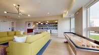 This single-floor amenity space showcases a cohesive mid-century modern aesthetic, unified by light wood-look tile flooring, textured geometric wallcoverings in warm gold tones, and a carefully curated color palette of mustard yellow, crimson red, and crisp white throughout.

The main lounge area serves as the heart of the space, anchored by a pair of plush mustard-yellow sofas arranged around a wall-mounted flat-screen television. An articulated black sputnik-style chandelier overhead adds a retro design touch, while recessed lighting ensures the space remains bright and welcoming.

Adjacent to the lounge, a game zone houses a substantial shuffleboard table positioned beneath large windows that frame open outdoor views, filling the area with abundant natural light. A separate dining nook features a round black pedestal table surrounded by cognac leather chairs, positioned beside expansive windows overlooking a lush green landscape.

The wet bar area is defined by white cabinetry, open shelving with patterned tile backsplash, globe pendant lights, and a built-in banquette upholstered in rich red velvet. Colorful abstract artwork and lush tropical plants are placed throughout, contributing to the space's vibrant, social atmosphere.