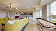 This single-floor amenity space showcases a cohesive mid-century modern aesthetic, unified by light wood-look tile flooring, textured geometric wallcoverings in warm gold tones, and a carefully curated color palette of mustard yellow, crimson red, and crisp white throughout.

The main lounge area serves as the heart of the space, anchored by a pair of plush mustard-yellow sofas arranged around a wall-mounted flat-screen television. An articulated black sputnik-style chandelier overhead adds a retro design touch, while recessed lighting ensures the space remains bright and welcoming.

Adjacent to the lounge, a game zone houses a substantial shuffleboard table positioned beneath large windows that frame open outdoor views, filling the area with abundant natural light. A separate dining nook features a round black pedestal table surrounded by cognac leather chairs, positioned beside expansive windows overlooking a lush green landscape.

The wet bar area is defined by white cabinetry, open shelving with patterned tile backsplash, globe pendant lights, and a built-in banquette upholstered in rich red velvet. Colorful abstract artwork and lush tropical plants are placed throughout, contributing to the space's vibrant, social atmosphere.