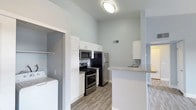 This single-story space showcases a well-appointed kitchen that serves as a central highlight. The kitchen features tall white shaker-style cabinetry that extends toward notably high ceilings, maximizing vertical storage. Stainless steel appliances include a gas range, built-in microwave, and a full-size refrigerator, complemented by a mosaic tile backsplash that adds a subtle decorative touch. A peninsula with a granite-style countertop provides additional workspace and casual seating potential, while a secondary sink is visible at the far end, suggesting added kitchen functionality. The flooring throughout is a wood-look plank in a neutral gray-toned finish, contributing to the cohesive, contemporary aesthetic. Walls are painted in a soft, light gray palette that enhances the airy feel created by the elevated ceilings. Adjacent to the kitchen, a dedicated laundry area is visible, housing a full-size washer and offering shelving for storage. Recessed and flush-mount ceiling lighting provides consistent illumination. The overall design is clean, functional, and modern.