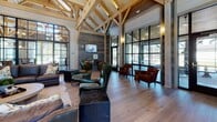 Located at 11000 Eagle Hall Ln in Goose Creek, South Carolina, this single-floor community clubhouse showcases an impressive rustic-modern design rooted in timber-frame craftsmanship. The centerpiece of the main gathering area is a dramatic vaulted ceiling supported by exposed natural wood trusses, lending the space a grand, barn-inspired character. Wide-plank hardwood flooring runs throughout, tying together the various seating areas with warmth and continuity.

A striking floor-to-ceiling stone fireplace anchors one end of the main lounge, paired with rich leather seating and layered textiles for a welcoming, lodge-like atmosphere. Shiplap and reclaimed wood wall paneling reinforce the rustic aesthetic while large, black-framed arched and grid windows flood the interior with abundant natural light and offer views of the surrounding landscape.

A dedicated seating alcove provides a quieter retreat, furnished with swivel accent chairs and wall-mounted media. The full kitchen features light wood cabinetry, dark countertops, open shelving, stainless steel appliances, and timber framing details, making it well-suited for catering and events.

The spacious screened porch extends the living area outdoors, finished with a wood-paneled ceiling, large tile flooring, and panoramic views of the grounds — ideal for year-round relaxation and social gatherings.