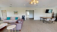 This clubhouse offers a well-organized, single-floor layout designed for community gathering and relaxation. The combined dining and living room serves as the heart of the space, featuring plush carpeted flooring, ceiling fans, and an abundance of natural light streaming through multiple windows. Multiple seating arrangements — including sofas, armchairs, and dining tables — provide flexible options for both casual lounging and group gatherings. A wall-mounted television adds a modern entertainment element, while a classic fireplace with a white surround and a rich brown accent wall creates a warm, inviting focal point in the living area. The kitchen is a functional galley-style space finished with warm wood cabinetry, dark countertops, and hardwood-style flooring. Stainless steel appliances — including a refrigerator, range, and microwave — equip the kitchen for community use. A connecting hallway links the kitchen to the main living areas, ensuring smooth flow throughout the space. A staircase provides additional access within the building. Overall, the clubhouse presents a bright, neutral palette with warm accents, combining practical amenities with a comfortable, community-focused ambiance ideal for residents to gather and connect.