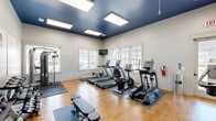 Located at 108 Cottage Creek Rd SW in Huntsville, Alabama, this single-floor fitness center offers a thoughtfully designed workout space with a generous footprint. The room's most distinctive architectural feature is its bold navy blue ceiling, accented by large recessed panel lighting that illuminates the space evenly and contributes to a modern, energetic ambiance. The white walls provide a clean contrast, while warm hardwood-style flooring runs throughout, adding a welcoming feel underfoot.

Multiple large windows line the walls, allowing abundant natural light to fill the space and providing views of the surrounding community, including a glimpse of an outdoor pool area. A wall-mounted flat-screen television is positioned near the cardio section for entertainment during workouts.

The fitness center is comprehensively equipped with TRUE-brand cardio machines, including treadmills, an elliptical, stationary bikes, and a rowing machine. A dedicated free weights area features a full dumbbell rack, kettlebells, and weight benches. A cable/multi-use strength machine is also available. Yoga-themed artwork adorns the walls, reinforcing the wellness-focused atmosphere. A posted fitness center rules board and fire safety equipment reflect attention to facility management and user safety.