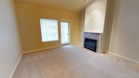 Located at 11400 Olympus Way in the charming waterfront city of Gig Harbor, Washington, Unit B302 is a thoughtfully designed single-floor residence that combines comfort and functionality. The living area serves as the heart of the space, featuring plush wall-to-wall carpeting that adds warmth underfoot. A tile-surround fireplace anchors the room, providing a cozy focal point ideal for the Pacific Northwest climate. Soaring walls with a tall chimney column lend a sense of vertical space and architectural interest to the interior. Natural light floods the room through a large window fitted with blinds, while a glass-paned door opens directly to a private balcony, seamlessly connecting the indoor living area to the outdoors. The warm, neutral yellow-toned walls create an inviting and bright atmosphere throughout. Track lighting overhead adds a practical and modern touch. The overall ambiance of the unit is clean, open, and move-in ready, making it an excellent choice for those seeking a comfortable residence in the Gig Harbor community.