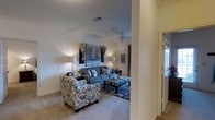 This single-floor residence offers a comfortable and cohesive living environment with a welcoming aesthetic throughout. The living area serves as the centerpiece of the home, featuring plush wall-to-wall carpeting in a warm neutral tone that adds softness underfoot and contributes to the room's inviting atmosphere. A ceiling fan is centrally mounted, providing both functional airflow and a classic finishing touch to the space. The room is furnished with a generously sized blue sofa accented with decorative pillows, a coordinating patterned armchair, and a striped ottoman that doubles as a coffee table — all arranged atop a detailed area rug that anchors the seating arrangement. Soft ambient lighting from table lamps on either side of the seating area creates a warm and relaxed ambiance. Wall art and framed décor add personality to the neutral-toned walls, while architectural details such as a structural column subtly define the transition between spaces. A doorway with sidelights allows natural light to filter into the entry, brightening the overall interior. The layout flows naturally, making efficient use of the available space while maintaining an open and airy feel suitable for both everyday living and entertaining.