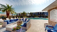 Located at 14801 Lemoyne Blvd in Biloxi, Mississippi, this single-level outdoor amenity space delivers a resort-style experience designed for relaxation and social gathering. At its center is a generously sized freeform swimming pool with clear turquoise water, complemented by an adjacent raised spa or water feature that adds a refined touch to the overall layout.

The expansive concrete sun deck surrounding the pool provides ample room for rows of lounge chairs and chaise seating, ideal for sunbathing under the Gulf Coast sky. A dedicated lounge area anchored by a stone fire pit serves as a central gathering point, furnished with plush wicker sectional sofas and Adirondack chairs dressed in coordinating blue cushions.

A wood pergola structure along one side of the deck creates a shaded retreat, outfitted with additional seating for a more intimate setting. Teal market umbrellas and bistro-style furniture are distributed throughout for casual outdoor dining and shade.

Tropical palm trees and manicured shrubs frame the entire area, evoking a warm, coastal ambiance consistent with the Mississippi Gulf Coast setting. The brick clubhouse building adjacent to the pool features large windows and covered patio access, seamlessly connecting the indoor and outdoor spaces. The surrounding multi-story residential buildings in a classic blue and gray palette complete the cohesive community aesthetic.
