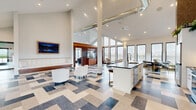 Located at 5 Ledge Dr in Schenectady, NY, this single-floor space offers a thoughtfully designed open-concept layout combining a kitchen and living area. The architecture is defined by dramatic vaulted ceilings that rise to impressive heights, creating a sense of grandeur and openness throughout. Exposed industrial ductwork adds a contemporary edge to the design.

The living area is anchored by an eye-catching geometric patterned floor featuring a blend of blue, gray, and neutral tones. Floor-to-ceiling windows line the walls, flooding the interior with abundant natural light while providing views of the surrounding neighborhood. A sliding glass door offers direct access to an outdoor patio area. Recessed lighting and wall-mounted sconces complement the natural light throughout.

The kitchen features white cabinetry, dark countertops, stainless steel appliances, and a large central island with an undermount sink. A textured accent wall adds warmth and visual interest. The open flow between the kitchen and living space makes the layout highly functional. Overall, the space presents a clean, modern aesthetic with a sophisticated and welcoming ambiance.