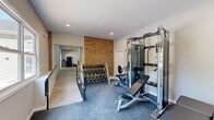 Located in Hickory, NC, this dedicated exercise room offers a well-appointed fitness facility designed to support a variety of workout routines. The space is defined by a striking exposed brick accent wall that adds an industrial-chic character, contrasting warmly with the clean white walls and recessed lighting above. The room is finished with durable grey carpeted flooring throughout, providing cushioning and traction underfoot. A large multi-panel window along one wall floods the space with natural light and offers pleasant views of the surrounding greenery and outdoor amenity areas, creating an inviting atmosphere for exercise. A glass exterior door provides direct outdoor access to a balcony or deck area, further enhancing the sense of openness. The fitness room is outfitted with a comprehensive selection of equipment, including a multi-function cable machine, an adjustable weight bench, a recumbent exercise bike, a treadmill, and a full rack of dumbbells spanning a wide range of weights. Recessed ceiling lights ensure bright, even illumination throughout the space. A metal railing near the entrance adds a modern architectural detail. This exercise room delivers a complete and functional fitness experience in a compact, well-designed setting.