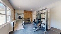 Located in Hickory, NC, this dedicated exercise room offers a well-appointed fitness facility designed to support a variety of workout routines. The space is defined by a striking exposed brick accent wall that adds an industrial-chic character, contrasting warmly with the clean white walls and recessed lighting above. The room is finished with durable grey carpeted flooring throughout, providing cushioning and traction underfoot. A large multi-panel window along one wall floods the space with natural light and offers pleasant views of the surrounding greenery and outdoor amenity areas, creating an inviting atmosphere for exercise. A glass exterior door provides direct outdoor access to a balcony or deck area, further enhancing the sense of openness. The fitness room is outfitted with a comprehensive selection of equipment, including a multi-function cable machine, an adjustable weight bench, a recumbent exercise bike, a treadmill, and a full rack of dumbbells spanning a wide range of weights. Recessed ceiling lights ensure bright, even illumination throughout the space. A metal railing near the entrance adds a modern architectural detail. This exercise room delivers a complete and functional fitness experience in a compact, well-designed setting.