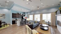 This single-story clubhouse offers a thoughtfully designed open-concept layout that seamlessly blends a fully equipped kitchen with a comfortable communal lounge area. The space is finished with warm hardwood-style flooring and soft blue-painted walls, creating a relaxed yet polished atmosphere throughout.

The kitchen area features dark espresso cabinetry, a black refrigerator, a dishwasher, and a countertop bar with stylish bar seating — ideal for casual gatherings. Pendant lighting hangs above the bar, complementing the recessed lighting embedded in the tray ceilings above.

The lounge area is generously sized and furnished with a plush gray sofa, accent armchairs in a bold yellow-and-white pattern, and a pair of round coffee tables. Twin ceiling fans help circulate air throughout the space. Large windows dressed with neutral curtains flood the interior with natural light while offering views of the surrounding outdoor area, enhancing the bright and airy ambiance.

Overall, this clubhouse strikes a balance between functional amenity space and inviting social environment, making it a versatile gathering place well-suited for both relaxed lounging and community events.