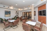 Located at 1409 Herrington Rd in Lawrenceville, Georgia, this single-floor communal lounge space offers a thoughtfully designed interior that blends modern sophistication with warm, inviting details. Light wood-look flooring runs throughout, complemented by paneled white walls adorned with curated artwork and decorative accents.

The main gathering area is furnished with multiple marble-topped tables surrounded by brown leather and gold-accented chairs, providing ample flexible seating for both casual meetings and social gatherings. A dedicated lounge nook features plush green upholstered armchairs arranged around a rustic wood coffee table, creating a comfortable retreat within the larger space.

A standout entertainment wall showcases a flat-screen TV framed by an exposed brick column and a rich wood-paneled accent surround, flanked by decorative botanical artwork. Stylish multi-arm track lighting adds a contemporary touch to the elevated ceilings throughout.

A wet bar area is set against a striking combination of red-painted brick and lighter stone tile accents, complete with dark countertops, a mini-refrigerator, and decorative vases. Large floor-to-ceiling windows flood the space with natural light while offering views of the surrounding exterior. Black-framed glass French doors connect to an adjacent room, enhancing the open, well-connected layout of this versatile communal environment.