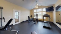 The Wales Crossing Fitness Center in North Canton, Ohio offers a comprehensively equipped workout facility designed to meet a variety of exercise needs. The single-floor space features vaulted ceilings that lend an open, airy feel, complemented by warm yellow walls and durable rubber flooring suited for high-traffic athletic use. A ceiling fan provides comfortable air circulation throughout the room.

Natural light streams in through large multi-pane windows along the main wall, offering pleasant outdoor views and creating a bright, energizing atmosphere. Wall-mounted flat-screen televisions are positioned for easy viewing during cardio sessions.

The cardio area is outfitted with a treadmill, an elliptical trainer, and a recumbent stationary bike, providing options for a range of aerobic workouts. A full-length mirror spans one wall, enhancing the sense of space while allowing users to monitor their form. The strength training section includes a multi-functional cable machine, an adjustable weight bench, and a well-stocked dumbbell rack with an organized selection of free weights. A glass-paned door connects the fitness room to an adjacent space, adding convenience and accessibility. Overall, the fitness center provides a clean, functional, and inviting environment for residents to maintain an active lifestyle.