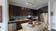 This single-floor space showcases a well-appointed modern kitchen designed with both aesthetics and functionality in mind. The kitchen is anchored by rich, dark espresso-toned cabinetry that extends from floor to ceiling, providing generous storage capacity throughout. The cabinetry is complemented by sleek brushed nickel hardware, contributing to the contemporary feel of the space.

Countertops are finished in light-toned granite, offering a striking contrast against the dark cabinets. A spacious kitchen island with a matching granite surface serves as a central focal point, ideal for meal preparation or casual dining. The backsplash features a mosaic tile design in subtle grey tones, adding texture and visual interest to the space.

The kitchen is fully equipped with stainless steel appliances, including a refrigerator, electric range, built-in microwave, and dishwasher. Overhead, a track lighting system provides targeted, adjustable illumination across the workspace. The overall ambiance is modern and polished, with a cohesive design palette that balances dark and light tones for an inviting yet sophisticated atmosphere.