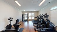 This single-floor fitness center is thoughtfully designed to support a full range of workout routines in a bright, clean environment. The space features warm hardwood flooring throughout, crisp white walls accented with crown molding, and recessed fluorescent lighting that ensures consistent illumination across the entire area.

Cardio equipment is well-represented, with a row of True-brand treadmills and elliptical trainers positioned along one wall, offering users a variety of aerobic options. Complementing these are multiple True recumbent stationary bikes, ideal for low-impact cycling workouts. A rowing machine rounds out the cardio selection, providing a full-body alternative.

Strength and functional training needs are addressed by a multi-station cable machine, along with a well-stocked accessory rack holding medicine balls, resistance bands, and a variety of stability and exercise balls in multiple sizes. These elements make the space suitable for diverse fitness goals, from rehabilitation to high-intensity training.

Natural light enters through several windows fitted with blinds, including a glass-panel exterior door that provides both ventilation and outdoor views of the surrounding property. The layout is open and organized, allowing for comfortable movement between equipment stations. Overall, this fitness center delivers a practical, professional-grade workout environment with a clean and inviting ambiance.