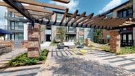 Located in Stafford, TX, this ground-floor unit is part of a well-appointed residential community that blends contemporary design with inviting outdoor living spaces. The exterior of the community showcases a mix of classic brick and clean horizontal lap siding, creating a refined architectural aesthetic that feels both modern and timeless.

The unit is accessible via a covered breezeway corridor framed by crisp white lap siding on both sides, lending a clean and well-maintained appearance. A metal staircase with sleek black railings leads to upper-level walkways, with an open second-floor balcony visible above, adding vertical dimension to the passage.

The standout feature of this community is the expansive landscaped courtyard, visible directly from the unit's entry. The courtyard is anchored by a striking wood-beam pergola supported by brick columns, beneath which a curated seating arrangement of modern lounge chairs and vibrant yellow accent tables creates an inviting outdoor social space. String lights overhead add ambiance, while mature trees, manicured greenery, and paved walkways complete the park-like setting.

The surrounding multi-story buildings frame the courtyard on multiple sides, creating a sense of enclosure and community. Open skies and lush landscaping contribute to a bright, airy atmosphere, making the outdoor spaces just as compelling as the interior living areas of this Stafford community.