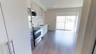This single-floor space showcases a clean, contemporary design with an open-concept layout that seamlessly connects the kitchen and living area. The kitchen is appointed with white cabinetry featuring sleek bar-pull hardware, light-toned countertops, and a full suite of stainless steel appliances, including a range with oven, built-in microwave, dishwasher, and refrigerator. A single-basin sink with a modern faucet is neatly integrated into the cabinetry along the wall. The kitchen flows naturally into an open living area finished with wide-plank, grey-toned hardwood-style flooring that runs throughout, lending a cohesive and contemporary feel to the space. Recessed lighting in the ceiling provides consistent illumination, complementing the abundant natural light that pours in through a large sliding glass door. This door opens directly onto a private balcony, offering outdoor access and views of the surrounding neighbourhood. The overall palette is neutral and bright, with white walls and light finishes creating a fresh, airy ambiance throughout the space.