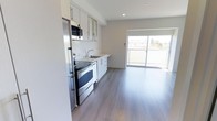 This single-floor space showcases a clean, contemporary design with an open-concept layout that seamlessly connects the kitchen and living area. The kitchen is appointed with white cabinetry featuring sleek bar-pull hardware, light-toned countertops, and a full suite of stainless steel appliances, including a range with oven, built-in microwave, dishwasher, and refrigerator. A single-basin sink with a modern faucet is neatly integrated into the cabinetry along the wall. The kitchen flows naturally into an open living area finished with wide-plank, grey-toned hardwood-style flooring that runs throughout, lending a cohesive and contemporary feel to the space. Recessed lighting in the ceiling provides consistent illumination, complementing the abundant natural light that pours in through a large sliding glass door. This door opens directly onto a private balcony, offering outdoor access and views of the surrounding neighbourhood. The overall palette is neutral and bright, with white walls and light finishes creating a fresh, airy ambiance throughout the space.