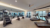 Located at 2925 Buford Dr in Buford, GA, this well-appointed fitness center offers a comprehensive workout environment designed with both functionality and aesthetics in mind. The single-floor facility features a spacious, open layout with neutral-toned carpet flooring laid in a herringbone pattern, recessed lighting, and crown molding details that lend a polished, upscale feel to the space.

The cardio zone is generously equipped with multiple treadmills — including a curved-deck model — stationary bikes, air bikes, elliptical trainers, and a Matrix stair climber, catering to a wide range of fitness routines. A full wall of floor-to-ceiling mirrors, accented with warm LED cove lighting, lines one side of the main workout area, enhancing the sense of openness and allowing users to monitor their form. Ceiling fans with wood-toned blades contribute to comfort during intense workouts.

Large, multi-pane windows flood the space with natural light and provide views of the surrounding outdoor areas, including a pool visible from the elliptical section. A wall-mounted flat-screen TV is positioned for easy viewing, and a built-in shelving unit offers convenient storage. Glass partition walls connect distinct zones within the facility, maintaining visual openness while defining separate workout areas. The overall ambiance is clean, bright, and modern.