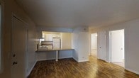 This single-floor unit offers a practical and well-organized layout designed for comfortable everyday living. The main living area features warm, rich hardwood-style flooring that adds character and a sense of coziness to the space. A defining feature of this unit is the pass-through kitchen, which connects seamlessly to the living area via a countertop bar — ideal for casual dining or food preparation. The kitchen is equipped with white cabinetry, a refrigerator, an electric range, and a sink, all framed within a compact and efficiently arranged footprint. The textured ceiling adds a subtle architectural detail overhead. Neutral, light gray walls throughout provide a versatile backdrop that complements a variety of interior styles. A doorway visible from the main living area leads to additional rooms, suggesting a thoughtful separation between living and private spaces. The lighter flooring in the adjoining room hints at a distinct material transition, contributing to a sense of spatial variety. Overall, the unit presents a clean, move-in-ready environment with a straightforward, functional design suited for a range of occupants.