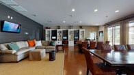 Located at 141 Hemlock Dr in Auburn, Alabama, this single-floor community clubhouse offers a thoughtfully designed gathering space that blends comfort with functionality. The interior is anchored by rich, polished concrete floors in a warm terracotta tone that flow seamlessly throughout the open layout, creating a cohesive and inviting atmosphere.

The lounge area serves as a central focal point, featuring a spacious sectional sofa accented with colorful throw pillows and a wall-mounted flat-screen TV set against a striking dark shiplap wood accent wall. A plaid upholstered ottoman and classic wooden dresser with table lamps add a warm, residential feel to the space.

Transitioning into the dining area, a large dark wood table surrounded by leather upholstered chairs provides ample seating for group gatherings. Banks of wide windows dressed with elegant drape panels flood the space with natural light and offer views of the surrounding exterior.

Along one wall, a row of built-in white shelving units with integrated desks and computer workstations provides a dedicated study and productivity zone, making the space equally suited for work and relaxation. Decorative accents, framed artwork, and curated accessories throughout contribute to the polished, welcoming character of this versatile community space.