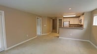 This single-floor space presents an open-concept layout that seamlessly connects the living area to the kitchen. The living space is finished with wall-to-wall carpeting in a neutral beige tone, complemented by warm tan walls and clean white baseboards, creating a cohesive and understated aesthetic throughout.

The kitchen is positioned along one side of the space and features white upper cabinetry, a stainless steel refrigerator, and a raised breakfast bar with a dark countertop that serves as a natural divider between the kitchen and living area. A utility sink is also visible, along with recessed and overhead lighting that illuminates the workspace effectively.

Recessed lighting in the ceiling provides focused illumination, while the overall layout feels open and functional. Several doorways lead to additional areas of the unit, including what appears to be a hallway with a built-in shelving unit, offering organizational storage options. A closet door is also visible along one wall. The single-floor design ensures convenient, step-free access throughout the entire space.