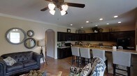 This two-story home showcases an open-concept layout on the main floor, where the kitchen, dining, and living areas flow together in a cohesive and functional arrangement. The kitchen is appointed with rich, dark cabinetry that extends to the upper walls, providing generous storage capacity. Granite countertops add a natural, textured elegance, and the space is anchored by a breakfast bar with upholstered bar seating — ideal for casual dining or entertaining. Recessed lighting throughout the kitchen ensures consistent, practical illumination, complemented by crown molding that adds a refined architectural detail along the ceiling line. The living area features polished concrete flooring in a warm, earthy tone, lending the space an industrial-chic character while remaining easy to maintain. A ceiling fan with a light fixture provides both air circulation and ambient lighting. A rounded archway connects the open living space to adjacent areas, adding a classic architectural touch. Decorative wall accents, including a sunburst mirror, contribute to the space's curated aesthetic. The overall ambiance is warm and inviting, blending contemporary finishes with comfortable, casual living.