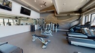 This modern fitness center is thoughtfully designed to support a comprehensive workout experience. The space is divided into distinct functional zones, each catering to different training needs. A standout feature is the dramatic accent wall finished in dark grey with flowing golden wave artwork, creating a dynamic and energizing atmosphere in the strength training area. This zone is outfitted with a cable machine, adjustable weight bench, dumbbells on a rack, a rowing machine, and additional resistance equipment, all set atop durable rubber flooring. A ceiling fan and wall-mounted flat-screen television add comfort and entertainment to this section. The cardio zone is positioned along a wall of large windows that frame scenic views of the surrounding landscape and an outdoor pool area, filling the space with natural light. Treadmills, a StairMaster, and elliptical machines are thoughtfully arranged to take full advantage of these views. High clerestory windows further enhance the sense of openness and airiness throughout the room. A dedicated stretching and functional training area provides ample open floor space, complemented by a storage rack holding exercise balls, resistance bands, and fitness accessories. Full-length mirrors are incorporated throughout, amplifying the sense of space and supporting proper form during workouts.