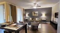 This single-floor space showcases a thoughtfully designed open-concept layout that seamlessly connects the dining and living areas. The living room is anchored by a warm, neutral-toned sofa accented with bold black-and-white striped throw pillows, complemented by a pair of cushioned accent chairs and a sleek glass-top coffee table. A striking charcoal-gray feature wall serves as a bold focal point, adorned with an expressive equestrian artwork that adds character and visual interest to the room. Warm hardwood flooring flows throughout the space, lending a sense of continuity and warmth. A ceiling fan with integrated lighting provides both comfort and ambient illumination, while flanking floor lamps cast a soft, inviting glow. Elegant crown molding adds a refined architectural touch to the ceiling line. Large windows dressed in rich brown drapes allow natural light to filter in while maintaining privacy. The adjacent dining area features a dark wood table set for entertaining, tying seamlessly into the overall sophisticated, earthy color palette. Together, the spaces create a welcoming and well-appointed environment ideal for both relaxing and hosting.