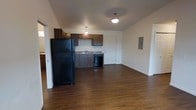 This single-floor residential unit offers a practical and functional layout centered around an open-concept living and kitchen area. The space is anchored by rich, dark hardwood flooring that extends throughout, providing a warm and cohesive aesthetic.

The kitchen is equipped with dark wood cabinetry offering ample storage, a granite-toned countertop, a stainless steel sink, a black refrigerator, and a black dishwasher. Pendant lighting hangs above, complementing the flush-mount ceiling fixture that illuminates the combined space. A utility panel is conveniently located along the wall near the kitchen, and a door provides additional access or storage off the kitchen area.

The open layout seamlessly connects the kitchen to the living area, creating a spacious and versatile environment suitable for everyday living. The neutral white walls allow for easy personalization, while the clean baseboards and simple design contribute to a tidy, move-in-ready appearance. The overall ambiance is functional and straightforward, offering a comfortable foundation for a well-organized living space.