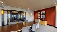 Situated in the heart of downtown Dallas at 400 N Ervay St, this professional office suite offers a polished and functional environment designed for executive meetings and collaborative work. The centerpiece of the space is a spacious conference room anchored by a large, dark-stained rectangular boardroom table surrounded by ergonomic upholstered chairs. A bold deep-red accent wall provides a striking visual contrast against the neutral tones of the surrounding walls and carpet, lending the room a sense of sophistication and energy. A gallery of framed black-and-white photographs adorns the accent wall, adding a curated, professional aesthetic. Pendant lights suspended from a ceiling-mounted track system cast warm, focused illumination throughout the room. A bank of large windows along one side of the conference room frames panoramic views of the Dallas skyline, flooding the space with natural light. Built-in cabinetry offers convenient storage and display surfaces. An adjacent support area features light hardwood flooring, dark-trimmed louvered doors, and a built-in workstation with office equipment, providing a practical complement to the main meeting space. Glass-paneled doors connect the rooms, maintaining openness while allowing for separation when needed.