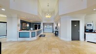 This sophisticated lobby presents a polished, welcoming environment designed to impress from the moment of arrival. The grand entrance is defined by soaring double-height ceilings adorned with detailed crown molding and a striking chandelier that anchors the space with warm, ambient light. Recessed lighting throughout complements the natural light streaming in through large glass entry doors and windows that overlook the exterior streetscape.

The reception area features a curved, teal-paneled desk positioned prominently near the entrance, providing an organized and professional check-in experience. Underfoot, large-format travertine tile in warm honey tones flows seamlessly across the entire space, enhancing the sense of openness and continuity.

Adjacent to the reception, a comfortable lounge area invites guests to relax beside a stone-surround fireplace, which serves as a refined architectural focal point. The lounge is thoughtfully furnished with a dark sofa, a teal wingback chair, and a rounded ottoman, creating an inviting conversational arrangement. A beverage station with a white cabinet and coffee service provides added convenience for visitors.

Decorative wall sconces, ornate mirrors, and framed artwork contribute to the space's refined aesthetic, while wide, well-lit corridors connect to additional areas beyond, ensuring smooth and intuitive circulation throughout.