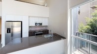 This two-story space showcases a sleek, contemporary design with clean lines and a bright, open aesthetic. The kitchen is a standout feature, boasting soaring double-height ceilings that amplify the sense of space. Crisp white cabinetry extends upward, complemented by dark wood-toned lower cabinets and a matching dark countertop that provides striking contrast. Stainless steel appliances, including a built-in microwave and a gas range, add a polished, functional touch. A peninsula with an undermount sink offers additional workspace and casual seating potential, while large windows flood the area with natural light, enhancing the airy atmosphere. The second floor is visible above, reinforcing the vertical openness of the layout. The outdoor balcony, enclosed by sleek glass-panel railings, provides an urban retreat with panoramic views of a vibrant city skyline. Mature treetops create a natural buffer, softening the cityscape below. This space combines modern interior design with an enviable outdoor connection to the surrounding urban environment.