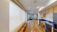 Located on S Mill St in the heart of downtown Jackson, Mississippi, this single-floor space offers a thoughtfully designed interior that balances comfort and functionality. Warm hardwood flooring runs throughout, complementing the clean white walls and suspended tile ceilings equipped with recessed lighting that keep the space bright and welcoming.

The open layout flows seamlessly from one area to the next. A generously sized lounge area anchors the space, furnished with a large sectional sofa, round coffee tables, and a wall-mounted flat-screen television, making it ideal for relaxed gatherings. Framed artwork adorns the walls, adding color and visual interest. A pair of teal accent chairs and a side table create a comfortable secondary seating nook.

A dedicated dining area features a solid wood table with seating for four, positioned beside large windows fitted with roller shades that diffuse natural light while maintaining privacy. The kitchenette is well-equipped with light wood cabinetry, dark granite countertops, a double basin sink, a dishwasher, and a full-size refrigerator, providing practical amenities for extended use.

Along one wall, roller shades line a row of windows, with a built-in wooden bench beneath offering additional seating. The overall ambiance is clean, modern, and functional, making this space well-suited for community use, professional gatherings, or hospitality purposes.