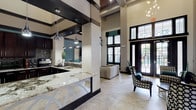 This well-appointed single-floor amenity space showcases a thoughtfully designed interior that blends contemporary style with functional versatility. The architecture is defined by dramatically soaring ceilings with bold dark trim banding, structural columns, and large black-framed windows and glass entry doors that flood the space with natural light and offer views of an outdoor pool area.

The main gathering area serves as the centerpiece, featuring large-format tile flooring, patterned lounge seating, and a wall-mounted flat-screen television set against a rich teal accent wall. A full kitchen is outfitted with dark cabinetry, mosaic tile backsplash, stainless steel appliances, and an expansive granite-topped bar with counter-height seating illuminated by sleek pendant lighting.

Adjacent to the main lounge, a game room accommodates a full-size billiards table alongside additional seating and another wall-mounted display. A separate, more intimate media room features warm hardwood flooring, teal-painted walls, a large mounted television, and stylish swivel chairs for comfortable viewing. A dedicated workstation area with individual computer terminals rounds out the space, offering a quiet zone for productivity within this multifaceted communal environment.