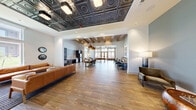 The Club Room at 150 Inspiration Dr in Hudson Oaks, Texas, is a thoughtfully designed community space that masterfully blends comfort and sophistication across a single, generously proportioned floor. The open-plan layout flows seamlessly between distinct functional zones, unified by warm hardwood flooring laid in a classic herringbone pattern throughout.

The lounge area near the entrance features a striking ornate tin ceiling with decorative pendant lighting, creating an immediate visual focal point. Comfortable leather seating and large windows invite natural light, offering pleasant views of the surrounding community grounds. The entry zone is anchored by steel-framed glass doors that add an industrial-chic contrast to the warm interior palette.

Deeper into the space, exposed wooden ceiling beams define a casual dining and gathering area furnished with a long communal table and mixed seating. A built-in banquette lined with upholstered bench seating, dark bistro tables, and decorative wall art creates an intimate café-style nook along one wall.

The kitchen area is equipped with stainless steel appliances, including a full-size refrigerator and under-counter unit, complemented by dark wood cabinetry and a patterned tile backsplash. Large exterior-facing French doors and clerestory windows flood the rear of the room with abundant natural light, completing this versatile and welcoming club room.