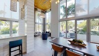 This thoughtfully designed single-floor space showcases dramatic double-height ceilings clad in warm wood paneling, creating an immediate sense of openness and architectural distinction. Towering floor-to-ceiling windows and expansive glass walls flood the interior with abundant natural light while framing lush views of tropical palm trees and outdoor seating areas beyond.

The space is organized into several distinct functional zones. A generous lounge area features plush gray sofas, a bold blue swivel accent chair, and an artfully layered area rug in blue and ivory tones, all anchored beneath sheer floor-length drapery panels that soften the natural light. Structural columns clad in white shiplap provide visual rhythm throughout the open plan.

A large dining area accommodates an extended dark wood table surrounded by bentwood chairs with patterned upholstery, positioned beneath a striking globe chandelier cluster and a soaring wood-paneled ceiling. Adjacent to the dining area, a well-appointed kitchenette features warm wood cabinetry, white quartz countertops, stainless steel appliances, and a vivid teal mosaic tile backsplash. Sculptural pendant lighting adds an artistic touch above the kitchen corridor, completing this polished, resort-inspired environment.