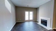 This single-floor space presents a well-appointed living area designed with both comfort and functionality in mind. The main living space is anchored by a tile-surround fireplace, offering a classic focal point that adds warmth and character to the room. Dark hardwood-style flooring runs throughout, providing a rich contrast against the neutral greige wall tones and crisp white baseboards and trim. A textured ceiling adds subtle architectural interest overhead, while a centrally placed HVAC vent ensures year-round climate control. A standout feature of the living area is the set of French doors, which flood the space with natural light and open to an outdoor deck or balcony, creating a seamless connection between interior and exterior living. The overall ambiance is clean, modern, and move-in ready, with a neutral color palette that offers a versatile backdrop for a variety of interior styles. The layout is open and unobstructed, allowing for flexible furniture arrangements and a sense of spaciousness. This space strikes a balance between understated elegance and everyday practicality, making it a welcoming and functional environment.