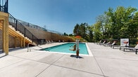 Located at 14735 Stable Gate Pl in Louisville, Kentucky, this two-floor outdoor amenity facility is centered around a generously sized swimming pool with crystal-clear water. The pool area is framed by a wide concrete sundeck furnished with modern lounge chairs and side tables, providing ample space for relaxation in the sun. Vibrant red patio umbrellas offer shaded seating areas equipped with tables and chairs for comfortable outdoor gatherings.

The facility features a two-story structure adjacent to the pool, with covered porches on both levels equipped with ceiling fans, offering a comfortable retreat from the sun. A wooden staircase with black metal railings connects the lower pool deck to the upper-level outdoor terrace, which overlooks the entire pool area. A stone retaining wall adds architectural character and defines the tiered layout of the space.

The pool area is fully enclosed by black metal fencing, ensuring safety while maintaining open sightlines to the surrounding mature tree canopy and green landscape. The natural backdrop of lush trees provides a serene and private atmosphere. The combination of functional amenities, thoughtful design, and scenic surroundings makes this a well-appointed outdoor recreational destination.
