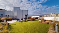 Located at 601 N Broad St in Philadelphia's vibrant Northern Liberties neighborhood, this expansive rooftop terrace offers a thoughtfully designed outdoor retreat above the city. The space is organized into distinct functional zones, seamlessly connected by wide stone pavers that guide visitors throughout the terrace. A lush artificial turf lawn serves as a central gathering area, flanked by lounge chairs ideal for relaxing and taking in the open sky. Surrounding the lawn, raised planting beds are filled with ornamental grasses, low-growing succulents, and seasonal shrubs, lending a naturalistic, garden-like character to the urban setting. Multiple seating clusters — including bistro-style table-and-chair sets and wicker lounge chairs — are positioned to take full advantage of the sweeping panoramic views of the Philadelphia skyline, visible in all directions beyond the clean white perimeter walls. The building's modern corrugated metal facade provides an architectural backdrop to the terrace, reinforcing the contemporary design aesthetic. Integrated bollard lighting ensures usability into the evening hours. The overall ambiance is one of urban sophistication balanced with green, open-air tranquility — a well-appointed outdoor amenity space designed for both relaxation and social gathering.