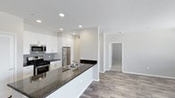 This single-floor space showcases a thoughtfully designed open-concept layout that connects the kitchen and living areas, creating a sense of spaciousness and easy flow throughout. The kitchen is a standout feature, equipped with stainless steel appliances—including a gas range, built-in microwave, and refrigerator—paired with crisp white cabinetry that extends to the ceiling for ample storage. A classic grey subway tile backsplash adds a refined touch, while the extended dark granite countertop peninsula doubles as a breakfast bar and provides generous workspace. A double-basin stainless steel sink is integrated into the peninsula, offering both functionality and a central focal point. Recessed lighting throughout the kitchen ensures a bright, clean ambiance. The living area continues the modern aesthetic with warm wood-look flooring that runs consistently through the space, enhancing visual continuity. Neutral, light-toned walls create a versatile backdrop suitable for a variety of interior styles. A hallway visible from the main living area leads to additional rooms, suggesting a practical and well-organized floor plan overall.