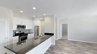 This single-floor space showcases a thoughtfully designed open-concept layout that connects the kitchen and living areas, creating a sense of spaciousness and easy flow throughout. The kitchen is a standout feature, equipped with stainless steel appliances—including a gas range, built-in microwave, and refrigerator—paired with crisp white cabinetry that extends to the ceiling for ample storage. A classic grey subway tile backsplash adds a refined touch, while the extended dark granite countertop peninsula doubles as a breakfast bar and provides generous workspace. A double-basin stainless steel sink is integrated into the peninsula, offering both functionality and a central focal point. Recessed lighting throughout the kitchen ensures a bright, clean ambiance. The living area continues the modern aesthetic with warm wood-look flooring that runs consistently through the space, enhancing visual continuity. Neutral, light-toned walls create a versatile backdrop suitable for a variety of interior styles. A hallway visible from the main living area leads to additional rooms, suggesting a practical and well-organized floor plan overall.