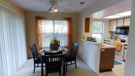 This single-story residence at Stonemill Village offers a welcoming and functional layout designed for comfortable everyday living. The dining area serves as a central gathering space, featuring a ceiling fan with integrated lighting overhead and a large window dressed with warm, sheer orange curtains that invite abundant natural light while offering a pleasant view of the surrounding greenery. The neutral-toned carpeting and light-colored walls create a bright, airy atmosphere throughout.

A defining architectural feature is the half-wall pass-through that connects the dining area to the kitchen, promoting an open flow between the two spaces. The kitchen, visible through the pass-through, includes raised cabinetry in warm wood tones and its own window, further enhancing the natural light within the home. The pass-through counter provides a convenient surface for casual use or display.

The overall ambiance of the space is cozy and inviting, with a layout that balances openness and defined zones. The single-floor design ensures ease of movement throughout, making it a practical and comfortable place to call home.