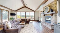 This generously proportioned single-floor space showcases a refined rustic-modern design that blends warmth and openness throughout. The most striking architectural feature is the dramatic vaulted ceiling with prominent exposed wood beam trusses, which soar overhead to create an expansive, airy atmosphere across the entire floor plan. Rich hardwood flooring runs throughout, complementing the natural wood tones of the beam work and door trim.

The living area serves as the heart of the space, anchored by a stone fireplace with a classic mantel that provides a cozy focal point. Large sectional seating and a patterned area rug define the lounge zone, while a wall-mounted television adds a modern functional touch. Adjacent to the living area, a dining space accommodates a full table and chair set, positioned near floor-to-ceiling windows that flood the room with natural light and frame verdant outdoor views.

A separate seating lounge area features neutral-toned armchairs arranged around a central table, ideal for intimate conversation. Oversized windows with warm wood framing appear throughout, connecting the interior to the surrounding landscape at every turn. Rustic wood-trimmed doorways and an entry corridor with framed artwork complete the inviting, cohesive aesthetic of this well-appointed space.