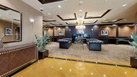 Located at 1026 S Broadway in the heart of Los Angeles, this elegantly appointed single-floor space offers a refined blend of luxury lounge and functional business center. The interior is defined by a striking coffered ceiling with recessed lighting and warm gold accent trim, lending an Art Deco-inspired grandeur to the room. Polished golden marble tile at the entry transitions seamlessly into plush neutral carpeting throughout the main lounge area.

The central seating arrangement features a pair of deep-buttoned, tufted black leather Chesterfield sofas flanking a sleek glass coffee table — an ideal setting for relaxed conversation or informal meetings. Framed black-and-white fine art photography adorns the dark accent walls, adding a cinematic, gallery-like atmosphere. Lush tropical plants positioned throughout the space provide natural contrast against the warm taupe and dark charcoal palette.

Along the perimeter, built-in workstations with ergonomic seating offer dedicated spaces for productivity, while a private nook furnished with plush orange armchairs and a side table provides a more intimate retreat. A large conference table in a secondary area accommodates formal meetings. Glass-paneled entry doors allow natural light to filter in, offering a glimpse of the outdoor greenery beyond. The overall ambiance is polished, professional, and distinctly upscale.