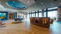 This striking single-floor amenity space exemplifies contemporary design at its finest, blending bold architectural elements with carefully curated interiors. The main lounge area commands attention with its dramatic teal-accented coffered ceiling, complete with a circular recessed feature fitted with ambient lighting that anchors the room's central gathering area. Light-toned large-format flooring runs throughout, reflecting the abundant natural light that streams in through floor-to-ceiling glass walls and expansive windows offering sweeping city and skyline views.

A curved wood reception desk with illuminated display niches serves as an elegant focal point near the entrance, showcasing decorative ceramic vessels. The lounge is furnished with a round, low-profile sofa set atop a golden area rug, complemented by a seating arrangement of modern reclining chairs with footrests accented in vibrant yellow.

Textural contrast is achieved through a sculptural 3D geometric wall panel and a dramatic marble-clad column, while deep teal accent walls are adorned with large-scale original artwork and a striking metallic wall installation near the elevator lobby. The space extends seamlessly outdoors to a furnished terrace equipped with a game table, outdoor seating, ceiling fans, and a mounted television, making it ideal for social gatherings and relaxation.