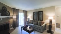 This single-floor space offers a warm and inviting atmosphere, centered around a thoughtfully designed living room. The room is anchored by a plush gray sofa adorned with an array of decorative pillows in neutral earth tones, complemented by a sleek dark-wood coffee table that adds a refined contrast to the light carpeted flooring. Natural light flows generously into the space through a large sliding glass door, which is elegantly framed by floor-to-ceiling patterned drapes in rich brown tones, adding depth and texture to the room. A coordinating table lamp provides soft ambient lighting, enhancing the relaxed yet polished feel of the space. The walls are finished in a warm beige tone, creating a neutral backdrop that highlights two framed botanical art pieces displayed prominently above the sofa. Subtle architectural details, including clean wall lines and white baseboards, contribute to the overall sense of understated elegance. The living area flows naturally, making it a comfortable and functional gathering space. The access to the outdoors via the sliding door suggests the potential for an adjacent patio or balcony, further extending the livable area and connection to natural surroundings.
