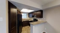 This single-floor space showcases a well-appointed kitchen as its central feature. The kitchen is designed with rich, dark espresso-toned cabinetry that extends from floor to ceiling, providing ample storage throughout. The countertops feature a granite-style surface in neutral earth tones, offering both visual appeal and practicality. A peninsula-style breakfast bar extends outward, creating a natural division between the kitchen and the adjoining living area while also offering additional counter space and seating potential.

Black appliances, including a refrigerator and an electric range with an overhead vent hood, complement the dark cabinetry for a cohesive aesthetic. A stainless undermount sink is positioned beneath a window that allows natural light to enter the space. A prominent skylight above the kitchen further enhances brightness, drawing in additional daylight and contributing to an open, airy feel.

The flooring throughout is wood-look laminate in a warm, natural tone, providing durability and a clean, contemporary look. The textured ceiling and neutral gray walls complete the overall design, creating a balanced and functional living environment.
