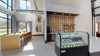 Located at 8801 Hidden River Pkwy in Tampa, FL, this single-story commercial space delivers a striking blend of modern industrial and contemporary design. The interior is defined by soaring double-height ceilings with exposed ductwork and bold circular pendant lighting, creating an open, airy atmosphere throughout.

The main lounge area features plush leather seating arrangements atop a dramatic dark area rug, anchored by a statement wall with an abstract marble-effect art installation. A dedicated workspace with a large communal table and colorful accent chairs adds functional versatility to the space.

A café-style service area serves as a visual centerpiece, highlighted by a vibrant geometric display wall in orange and black tones, refrigerated glass display cases, and a coffee station. An adjacent bar-height island with warm wood tones and orange stools offers additional seating.

Floor-to-ceiling windows and glass-paneled doors flood the interior with natural light while framing sweeping views of the lush, tree-lined landscape outside. Light grey large-format tile flooring flows seamlessly throughout, complementing the crisp white walls adorned with colorful abstract artwork.

A separate waiting or media area includes wall-mounted display screens and sleek metal seating. The grand entryway features double glass doors flanked by tall windows with elegant grey drapery, offering a welcoming and professional first impression.